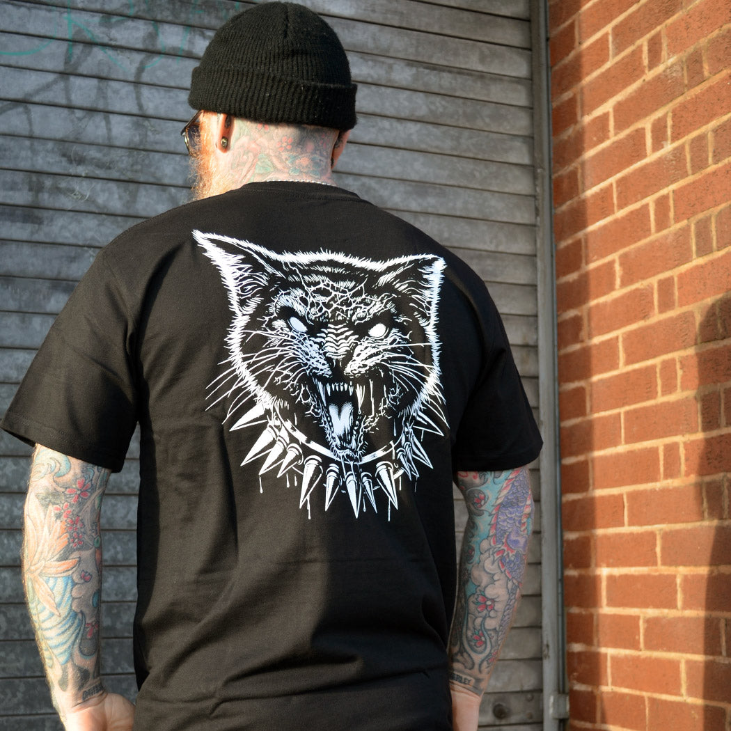 Person wearing a black t-shirt with a white graphic of a cat on the back, standing against a brick wall.
