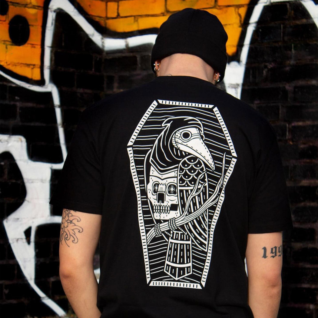 Person wearing a black t-shirt with a white graphic design on the back, standing in front of a graffiti-covered wall.