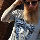 Man wearing a t-shirt with a black and white eye skull design against a brick wall.