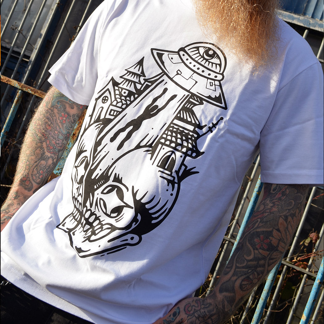 Person wearing a white t-shirt with a black graphic design, standing against a metal fence.