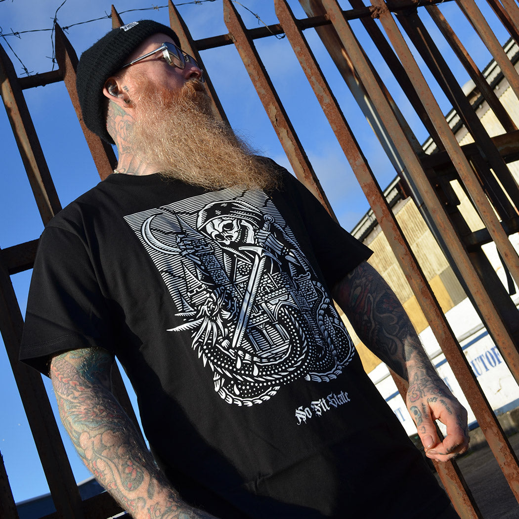Man wearing a black t-shirt with a medieval skull design, standing in front of a rusty metal fence.