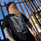 Man wearing a black t-shirt with a medieval skull design, standing in front of a rusty metal fence.