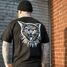 Person wearing a black t-shirt with a white graphic of a cat on the back, standing against a brick wall.