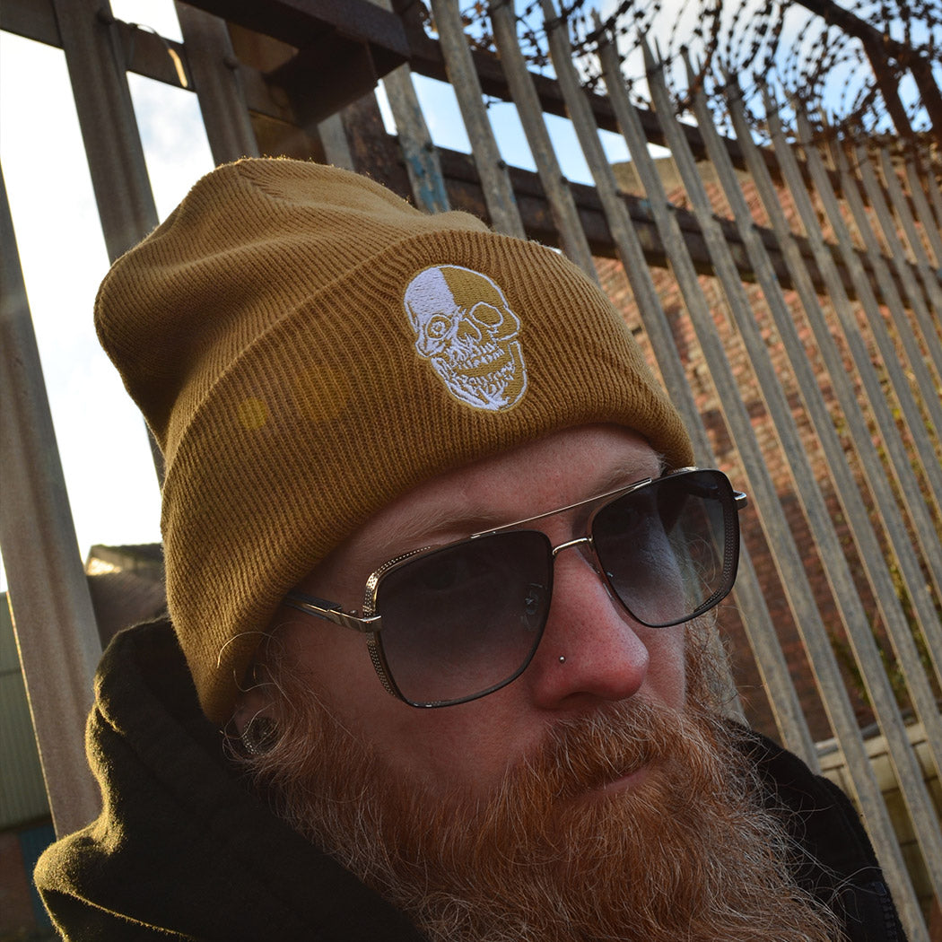 Person wearing a caramel coloured beanie with a skull design, standing in front of a metal fence.