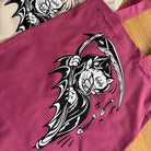 Burgundy tote bag with black and white graphic design of a grim reaper cat on a wooden surface.