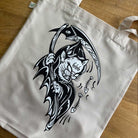Natural tote bag with black and white graphic design of a grim reaper cat on a wooden surface.